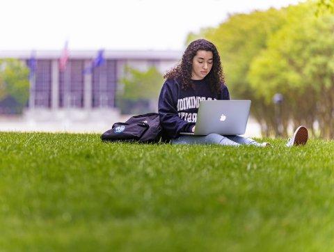 A female student wearing an ODU sweatshirt is studying on Kaufman Mall with her laptop resting on her legs